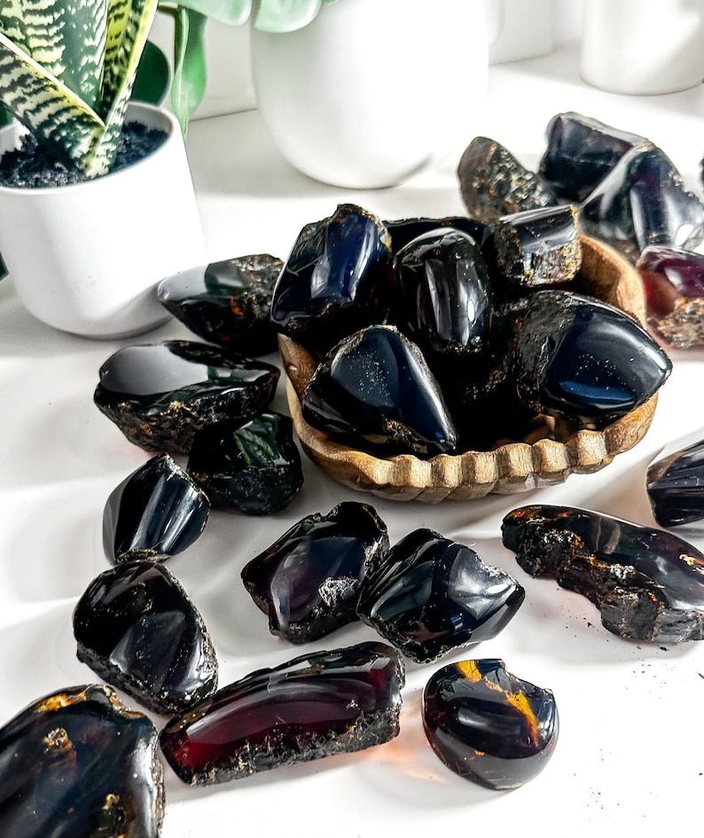 Puede incluir: Una colecci&oacute;n de piedras de &aacute;mbar oscuro, algunas con un tono rojizo, en varias formas y tama&ntilde;os. Algunas piedras est&aacute;n en un cuenco de madera, mientras que otras est&aacute;n esparcidas sobre una superficie blanca.