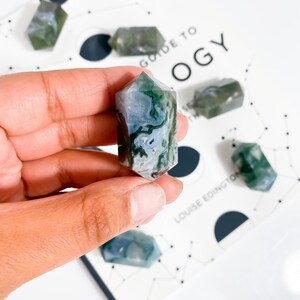 May include: A selection of moss agate crystals, with a focus on a hexagonal crystal held in a hand. The crystals display green and white patterns. The background includes a book with the text "Guide to OGY".
