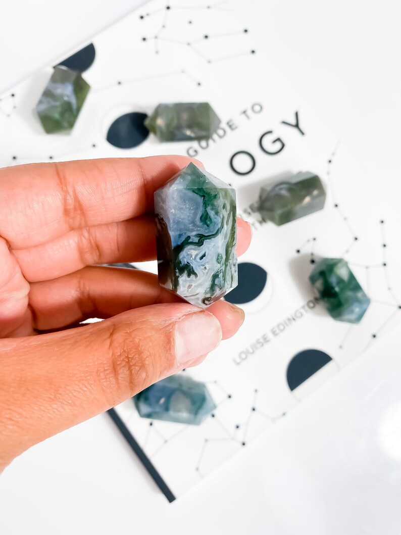 May include: A selection of moss agate crystals, with a focus on a hexagonal crystal held in a hand. The crystals display green and white patterns. The background includes a book with the text "Guide to OGY".