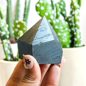 May include: A dark gray, faceted, geometric crystal in the shape of a pyramid. The crystal is being held in a hand with a floral design on the fingernail.