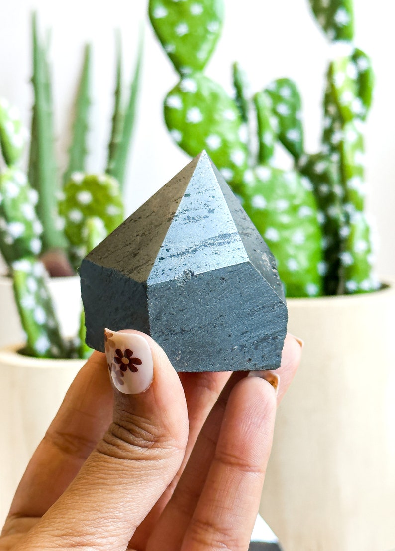 May include: A dark gray, faceted, geometric crystal in the shape of a pyramid. The crystal is being held in a hand with a floral design on the fingernail.