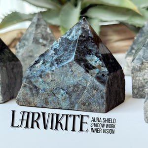 Pode incluir: Várias pirâmides de cristal de larvikite com iridescência cinza escuro e azul. Os cristais são facetados e têm uma textura áspera e natural. O texto na imagem diz "LARVIKITE" e "AURA SHIELD, SHADOW WORK, INNER VISION."