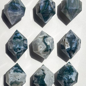 May include: Nine double-terminated moss agate crystals. Each crystal has a diamond-like shape with facets. The stones display a mix of translucent white, grey, and green hues, with moss-like inclusions. The crystals are arranged on a white surface.