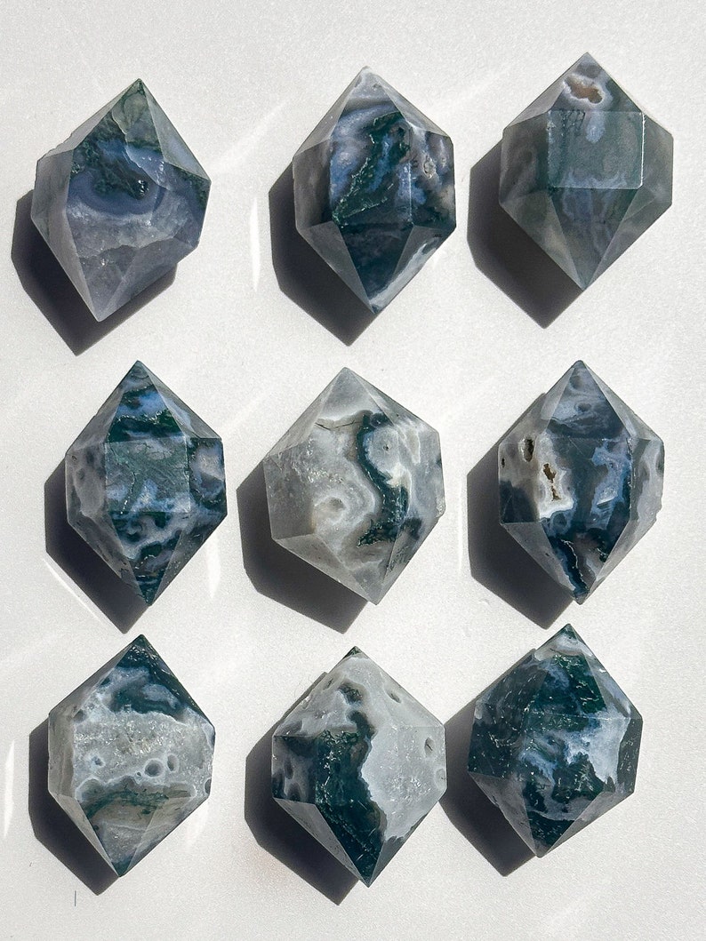 May include: Nine double-terminated moss agate crystals. Each crystal has a diamond-like shape with facets. The stones display a mix of translucent white, grey, and green hues, with moss-like inclusions. The crystals are arranged on a white surface.