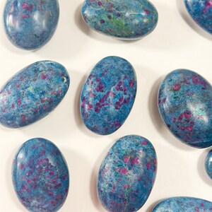 May include: A collection of polished, oval-shaped stones featuring a vibrant blue base colour with scattered, irregular magenta and deep pink inclusions throughout. Each stone has a smooth, glossy finish that reflects light across its rounded surface.