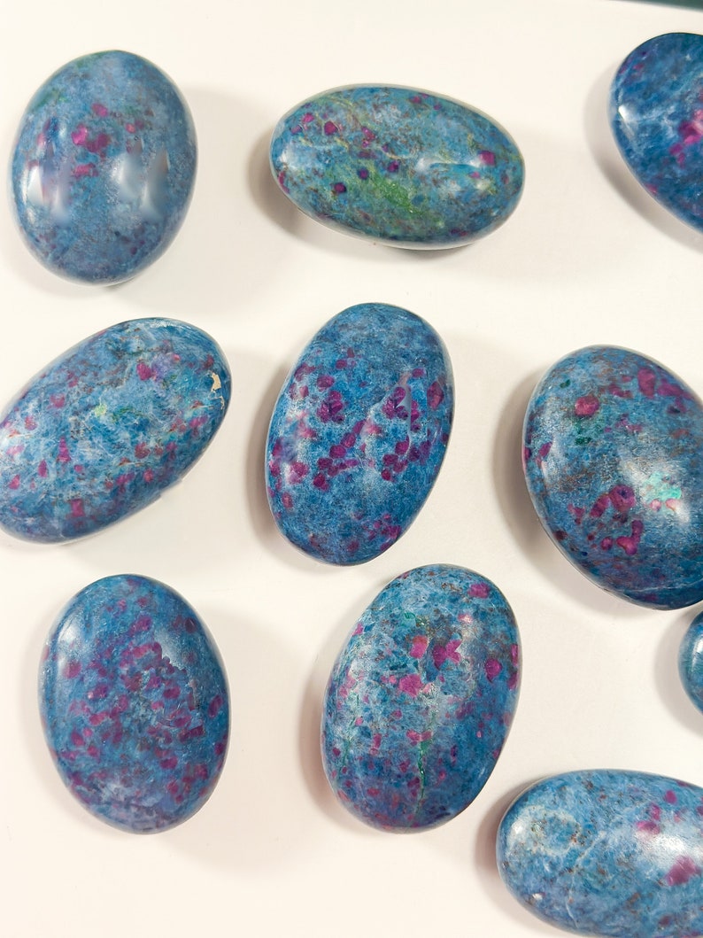 May include: A collection of polished, oval-shaped stones featuring a vibrant blue base colour with scattered, irregular magenta and deep pink inclusions throughout. Each stone has a smooth, glossy finish that reflects light across its rounded surface.