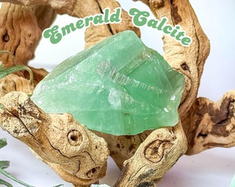 Puede incluir: Un gran cristal de calcita verde esmeralda translúcido se muestra sobre un fondo de madera flotante marrón claro y desgastada. El texto "Emerald Calcite" está escrito en una fuente verde caprichosa sobre el cristal.