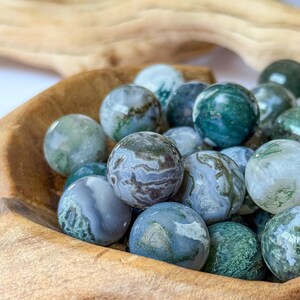 May include: A wooden bowl filled with polished, round gemstones. The stones vary in color, with shades of green, blue, and gray, and feature unique patterns. The bowl is made of light brown wood, and the background is blurred.