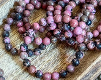 Puede incluir: Un conjunto de pulseras de piedras preciosas rosas y negras hechas de cuentas redondas. Las pulseras están apiladas sobre una superficie de madera.