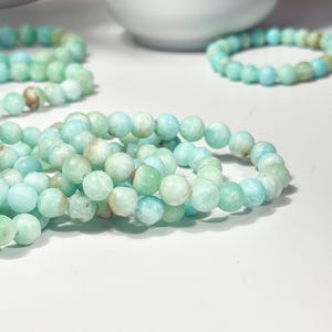 May include: Several strands of beaded bracelets. The bracelets are made of round, light blue and green beads. The beads have a slightly mottled appearance. The bracelets are arranged on a white surface, with a white ceramic object in the background.