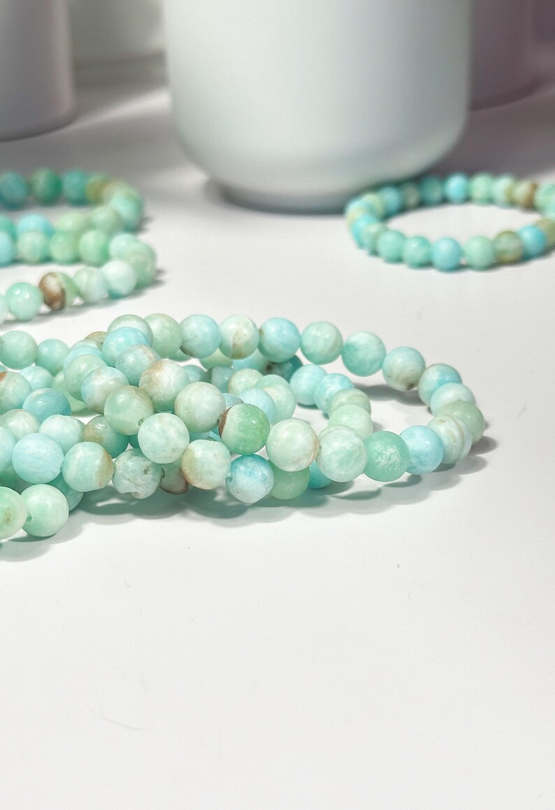 May include: Several strands of beaded bracelets. The bracelets are made of round, light blue and green beads. The beads have a slightly mottled appearance. The bracelets are arranged on a white surface, with a white ceramic object in the background.