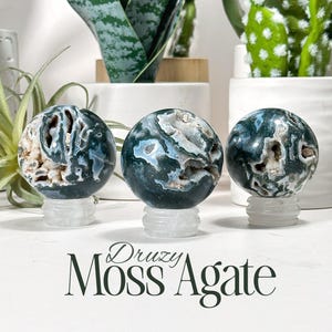 May include: Three spherical moss agate stones with visible druzy crystal inclusions, each resting on a clear circular stand. The stones feature deep green and white patterns. The text Druzy Moss Agate is printed in black at the bottom of the image.