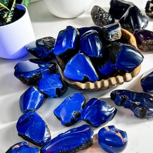 Puede incluir: Una colecci&oacute;n de piedras de &aacute;mbar azul, algunas en un cuenco de madera, otras esparcidas sobre una superficie blanca. Las piedras son de varios tama&ntilde;os y formas, con un color azul intenso y algunas inclusiones negras.