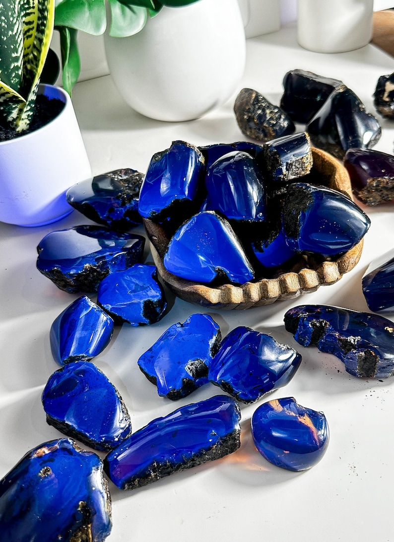 Puede incluir: Una colecci&oacute;n de piedras de &aacute;mbar azul, algunas en un cuenco de madera, otras esparcidas sobre una superficie blanca. Las piedras son de varios tama&ntilde;os y formas, con un color azul intenso y algunas inclusiones negras.