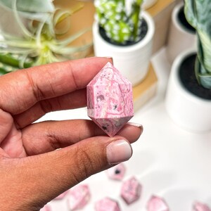 May include: A hand holds a pink and white double-terminated crystal point. The crystal has a hexagonal shape with a pointed end on each side. Other similar crystals are scattered in the background. The crystals are a light pink colour with white and black inclusions.