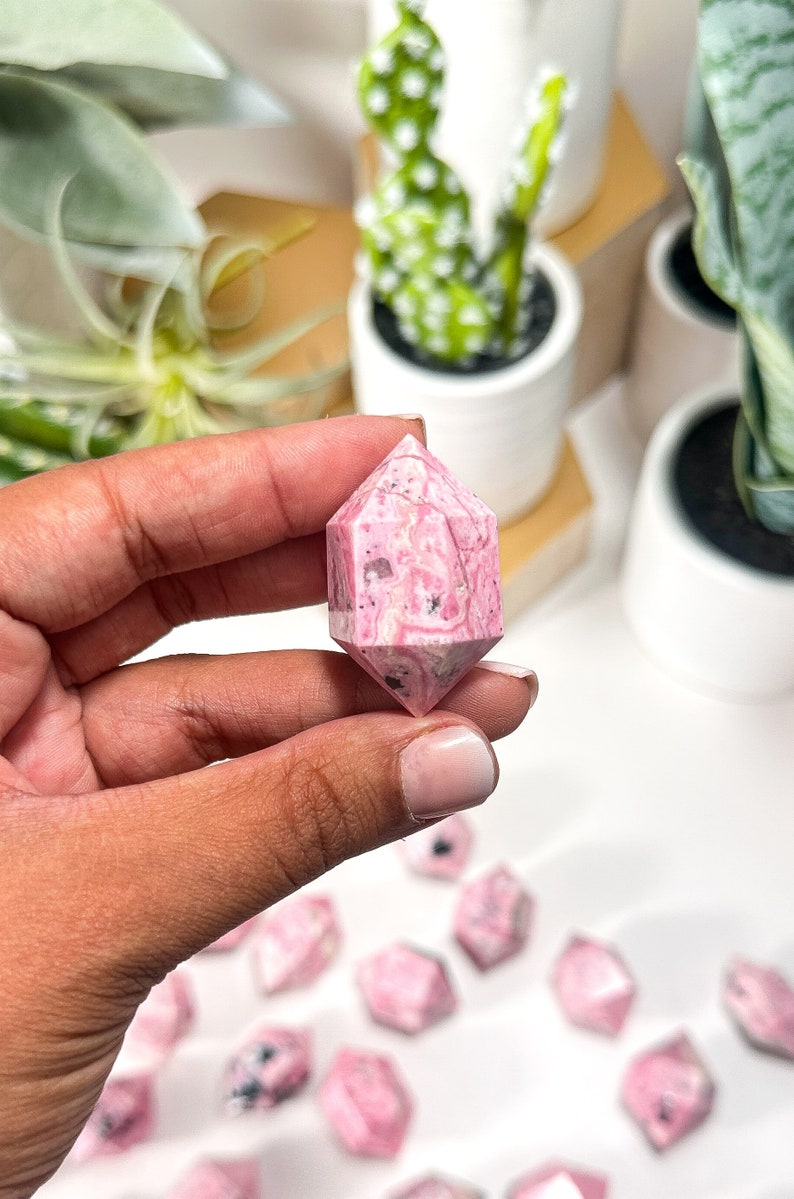 May include: A hand holds a pink and white double-terminated crystal point. The crystal has a hexagonal shape with a pointed end on each side. Other similar crystals are scattered in the background. The crystals are a light pink colour with white and black inclusions.