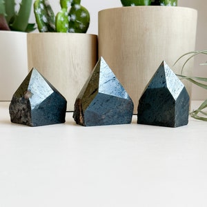 May include: Three dark gray, faceted, crystal points arranged in a row on a white surface. The points are all roughly the same size and shape.