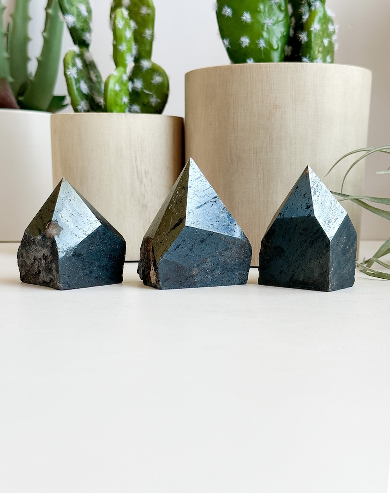 May include: Three dark gray, faceted, crystal points arranged in a row on a white surface. The points are all roughly the same size and shape.