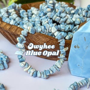 Pode incluir: Uma pulseira feita de pequenas pedras de Opala Azul Owyhee azul claro e branco. A pulseira é exibida em frente a uma tigela de madeira cheia de pedras semelhantes. O texto "Owyhee Blue Opal" é sobreposto na pulseira.