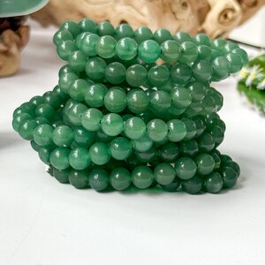 Puede incluir: Una pila de pulseras de cuentas verdes. Las cuentas redondas tienen un acabado pulido. Las pulseras est&aacute;n apiladas sobre una superficie blanca, con una escultura verde y madera flotante en el fondo.