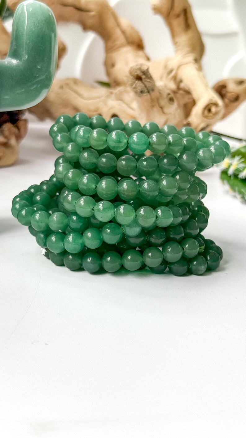 Puede incluir: Una pila de pulseras de cuentas verdes. Las cuentas redondas tienen un acabado pulido. Las pulseras est&aacute;n apiladas sobre una superficie blanca, con una escultura verde y madera flotante en el fondo.