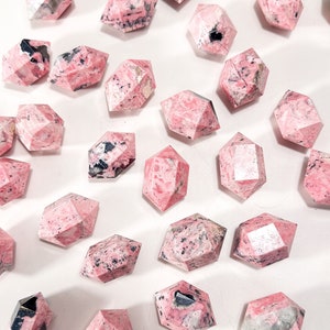May include: A selection of faceted, pink gemstone crystals with black and white inclusions. The crystals are in various shapes, including hexagonal and elongated forms. The gemstones are arranged on a white surface.