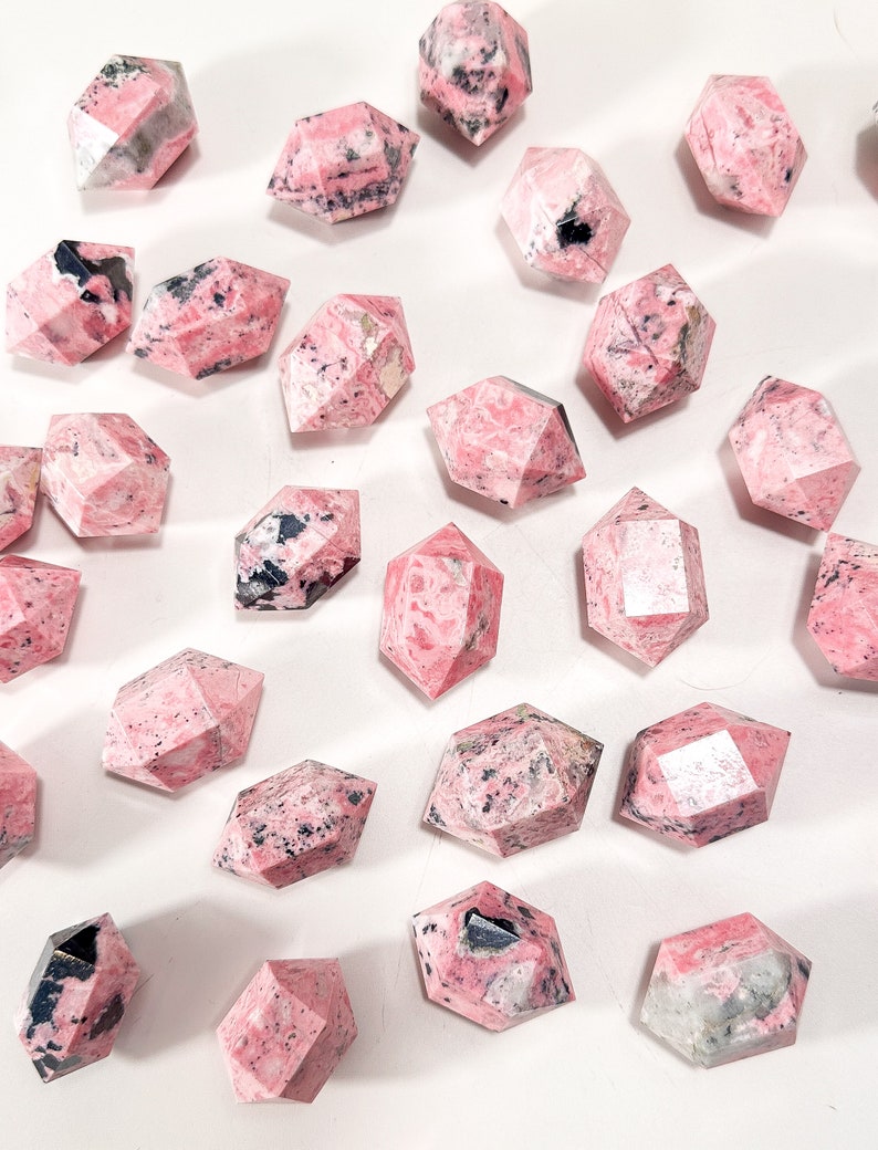 May include: A selection of faceted, pink gemstone crystals with black and white inclusions. The crystals are in various shapes, including hexagonal and elongated forms. The gemstones are arranged on a white surface.