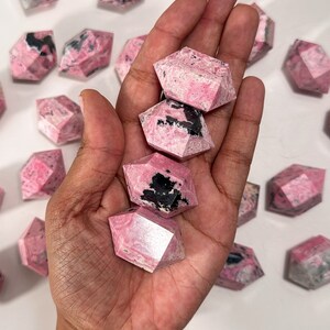 May include: A collection of pink and black rhodonite crystals. Several of the crystals are held in a hand, with others scattered around. The crystals have a geometric, faceted design and a mix of pink and black coloration.