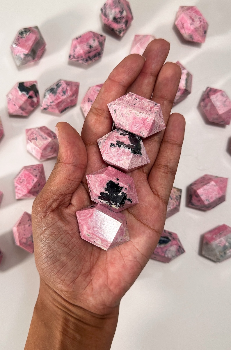 May include: A collection of pink and black rhodonite crystals. Several of the crystals are held in a hand, with others scattered around. The crystals have a geometric, faceted design and a mix of pink and black coloration.