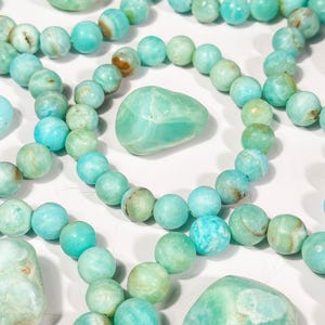 May include: An assortment of turquoise-colored beaded bracelets and polished stones. The beads and stones vary in shades of blue and green, with some showing brown inclusions. The bracelets are arranged in a circular pattern.