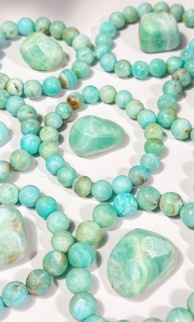 May include: An assortment of turquoise-colored beaded bracelets and polished stones. The beads and stones vary in shades of blue and green, with some showing brown inclusions. The bracelets are arranged in a circular pattern.