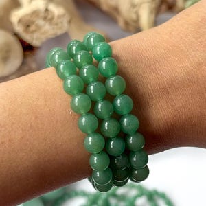 Puede incluir: Tres pulseras de piedras preciosas de aventurina verde. Cada pulsera est&aacute; hecha de cuentas redondas y pulidas. Las pulseras se muestran en un brazo, con otros productos de aventurina verde en el fondo.