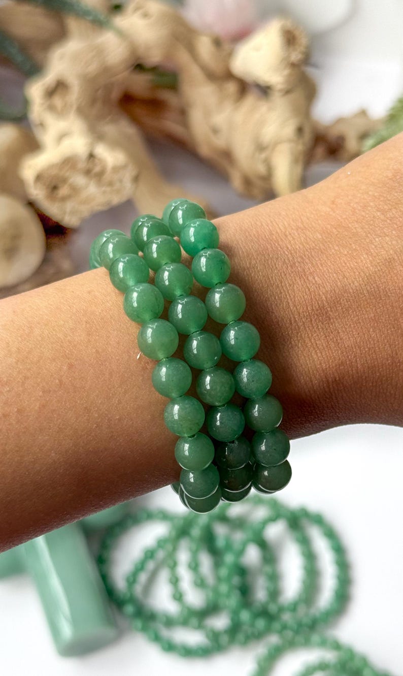 Puede incluir: Tres pulseras de piedras preciosas de aventurina verde. Cada pulsera est&aacute; hecha de cuentas redondas y pulidas. Las pulseras se muestran en un brazo, con otros productos de aventurina verde en el fondo.