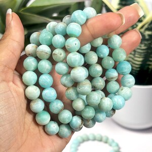 May include: A collection of round, light blue Amazonite gemstone beads. The beads are strung together to form bracelets. The bracelets are held in a hand, with a few more bracelets visible in the background.