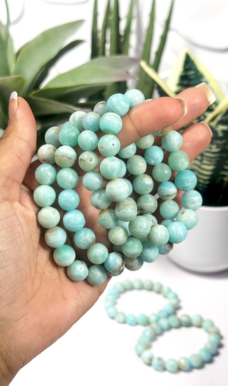 May include: A collection of round, light blue Amazonite gemstone beads. The beads are strung together to form bracelets. The bracelets are held in a hand, with a few more bracelets visible in the background.