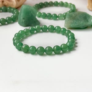Puede incluir: Primer plano de varias pulseras de jade verde con cuentas lisas y redondas. Las pulseras se exhiben sobre una superficie blanca, con algunas piedras de jade en bruto y detalles de madera en el fondo. Las cuentas son verdes.