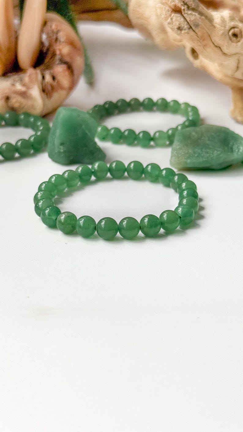 Puede incluir: Primer plano de varias pulseras de jade verde con cuentas lisas y redondas. Las pulseras se exhiben sobre una superficie blanca, con algunas piedras de jade en bruto y detalles de madera en el fondo. Las cuentas son verdes.