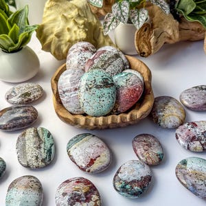 May include: A collection of polished, oval-shaped stones featuring a variety of speckled and marbled patterns in shades of mint green, deep red, brown, and white. Several stones are contained within a small, carved wooden bowl.
