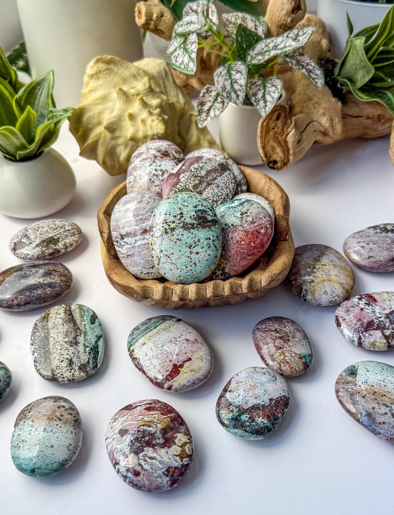May include: A collection of polished, oval-shaped stones featuring a variety of speckled and marbled patterns in shades of mint green, deep red, brown, and white. Several stones are contained within a small, carved wooden bowl.
