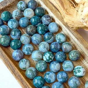 May include: A collection of polished moss agate spheres in varying shades of green, blue, and white, displayed in a wooden tray. The spheres are approximately 1 inch in diameter, showcasing natural patterns and textures. The arrangement highlights the natural beauty of the gemstones.