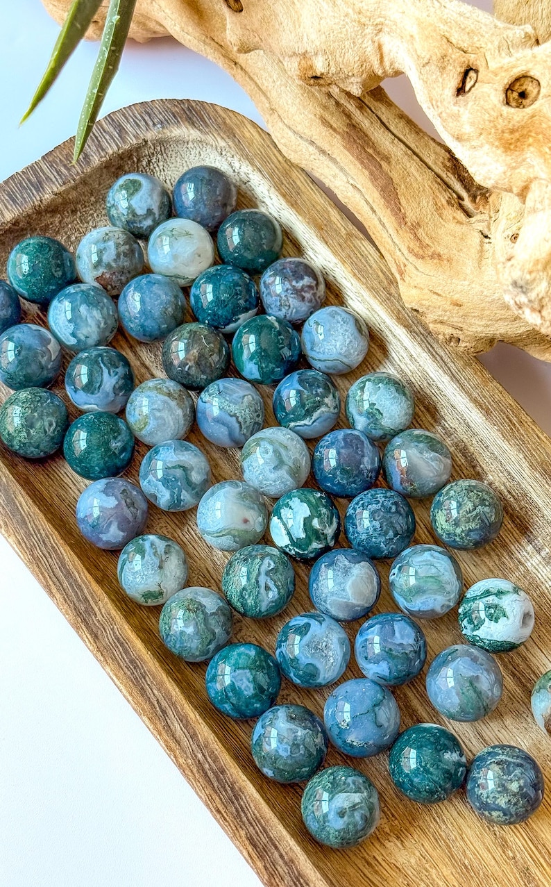 May include: A collection of polished moss agate spheres in varying shades of green, blue, and white, displayed in a wooden tray. The spheres are approximately 1 inch in diameter, showcasing natural patterns and textures. The arrangement highlights the natural beauty of the gemstones.