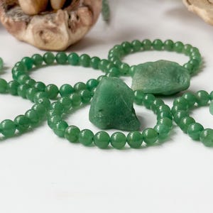 Puede incluir: Pulseras de aventurina verde y piezas de piedra en bruto se exhiben sobre una superficie blanca. Las pulseras est&aacute;n hechas de peque&ntilde;as cuentas redondas. Las piedras en bruto tienen una textura rugosa y natural. El fondo incluye elementos de madera y un hongo.