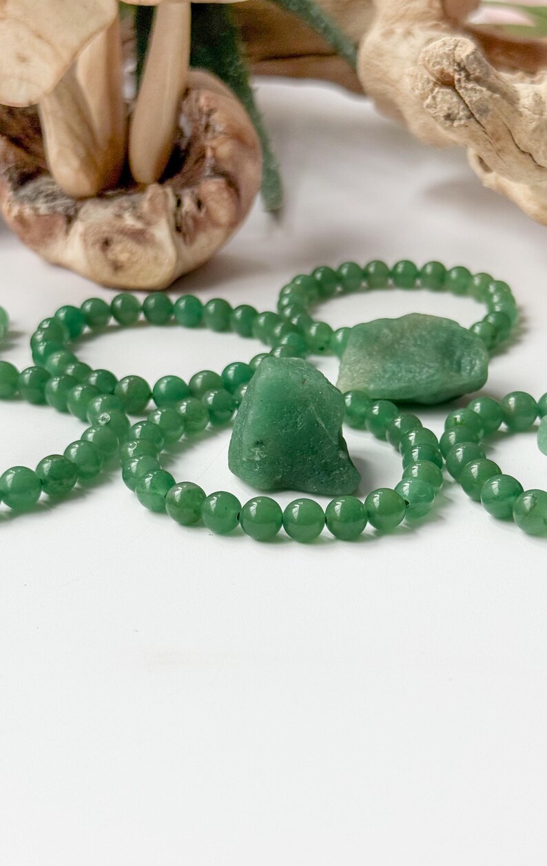 Puede incluir: Pulseras de aventurina verde y piezas de piedra en bruto se exhiben sobre una superficie blanca. Las pulseras est&aacute;n hechas de peque&ntilde;as cuentas redondas. Las piedras en bruto tienen una textura rugosa y natural. El fondo incluye elementos de madera y un hongo.