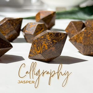 May include: Close-up of several faceted jasper stones with intricate brown and gold patterns. The stones are arranged on a white surface. The word "Calligraphy" and the word "Jasper" are written in a cursive font.