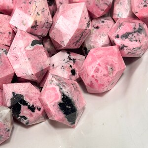 May include: A pile of pink gemstones with a faceted design and black and white markings. The stones are of varying sizes and shapes, with a geometric cut. The image highlights the natural patterns and textures of the mineral specimens.