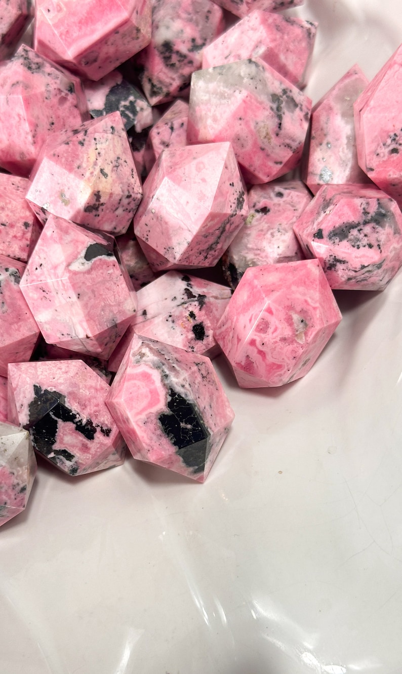 May include: A pile of pink gemstones with a faceted design and black and white markings. The stones are of varying sizes and shapes, with a geometric cut. The image highlights the natural patterns and textures of the mineral specimens.