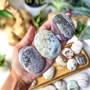 May include: Three smooth, oval-shaped polished stones held in a palm. The top stone is white with green and maroon patches. The middle stone is cream-coloured with dark speckles and a small geode centre. The bottom stone is mauve with dark spots.
