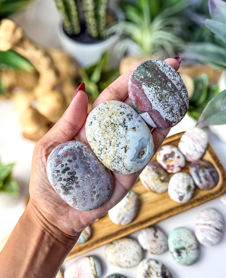 May include: Three smooth, oval-shaped polished stones held in a palm. The top stone is white with green and maroon patches. The middle stone is cream-coloured with dark speckles and a small geode centre. The bottom stone is mauve with dark spots.