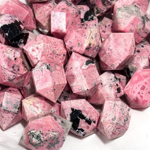 May include: A pile of faceted, pink rhodonite crystals with black markings. The crystals are geometrically shaped and presented in a white container. The pink hue varies, offering a visually interesting arrangement.