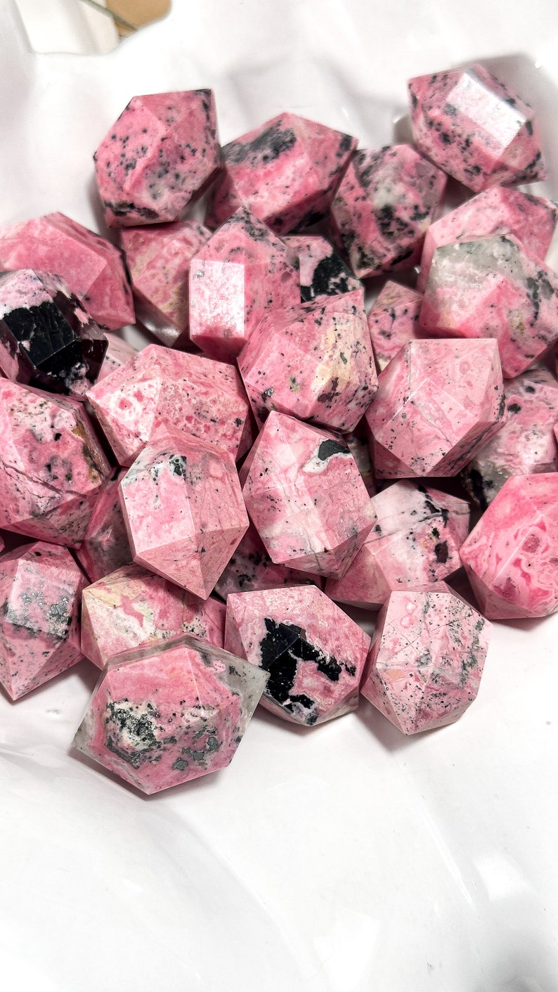 May include: A pile of faceted, pink rhodonite crystals with black markings. The crystals are geometrically shaped and presented in a white container. The pink hue varies, offering a visually interesting arrangement.
